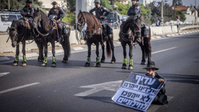מפגין מהפלג הירושלמי יושב על הכביש עם שלט נגד חוק הגיוס ופרשי משטרה ברקע